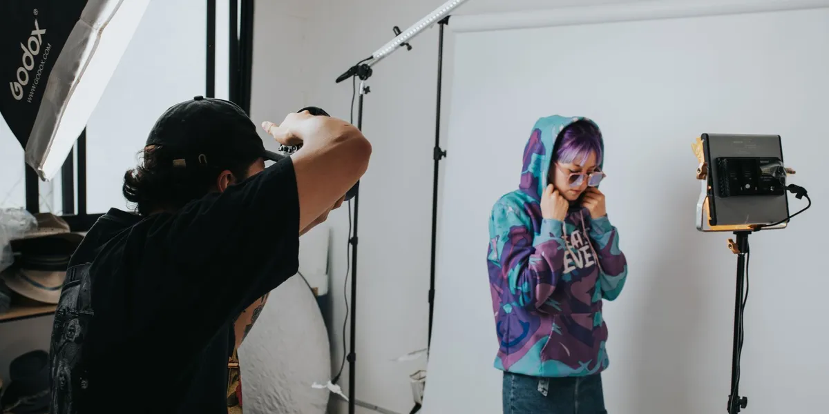 A person stands in front of a white paper backdrop lit by an LED panel on a stand, a large softbox and an overhead LED light bar on a boom, while a camera operator frames the shot.