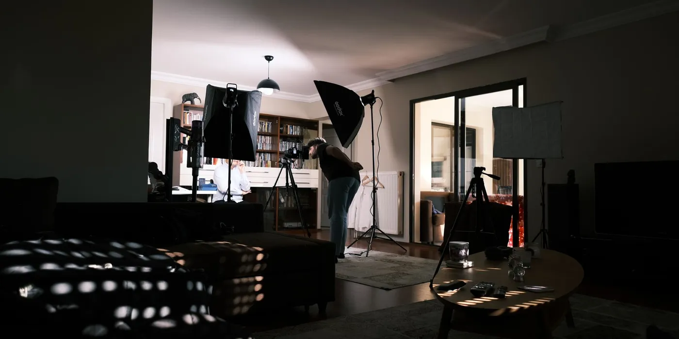 A living room converted into a small interview set, with two softboxes on stands, a camera on a tripod and an operator leaning in to check the framing on a seated subject.